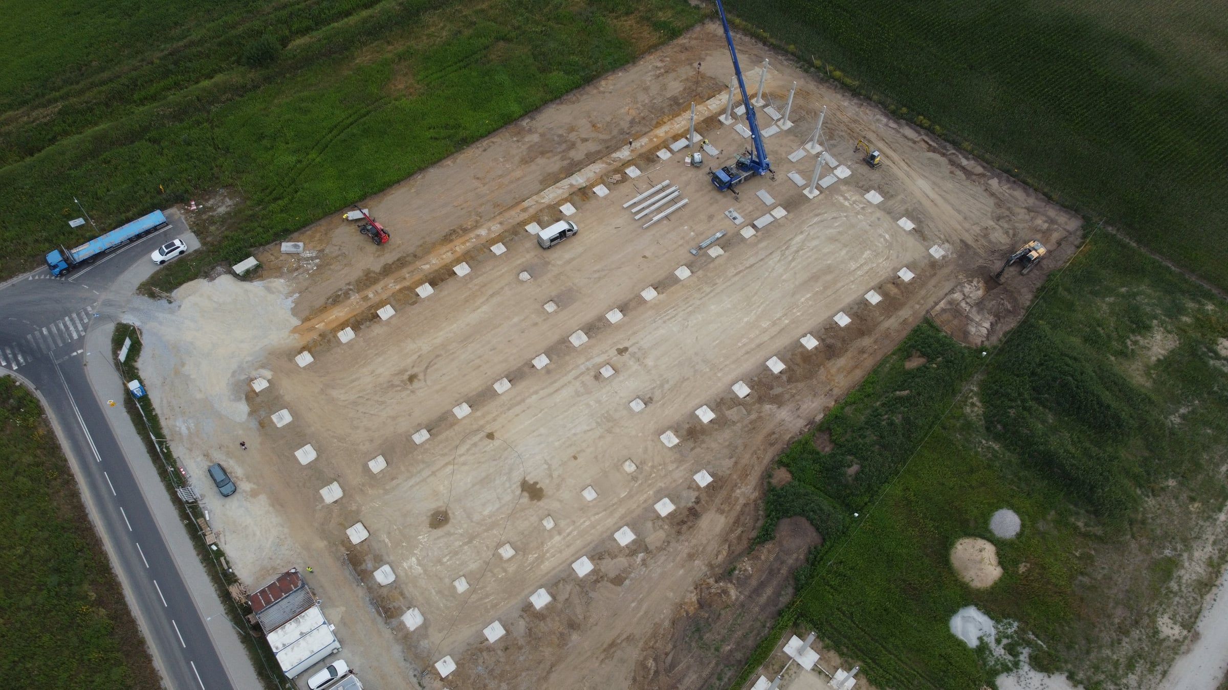Widok z drona na cały plac budowy: rzędy gotowych stóp fundamentowych na obrysie hali i dźwig przy pierwszych słupach.