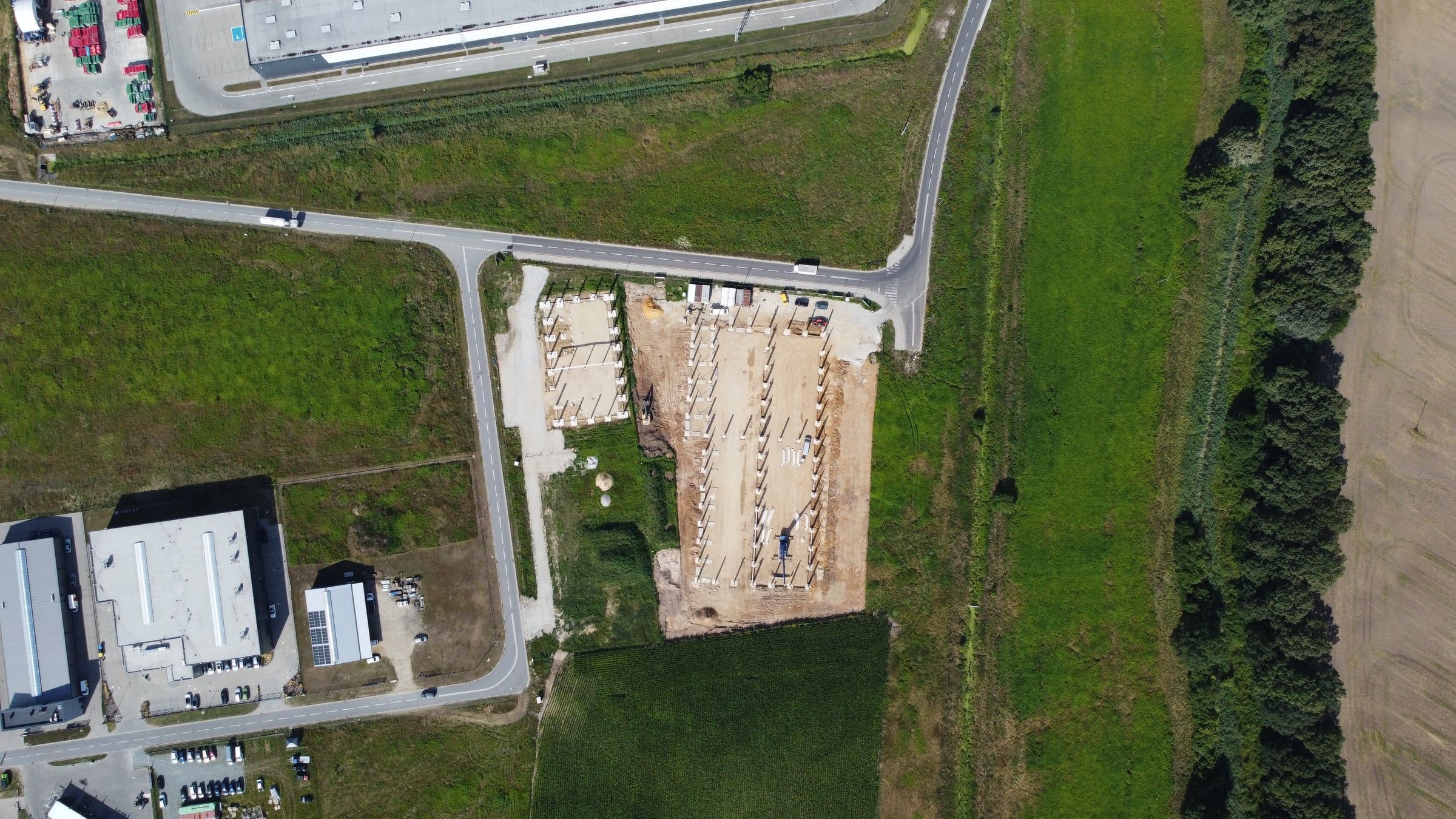 Widok z dużej wysokości na budowę na tle okolicy: plac budowy hali, drogi dojazdowe i sąsiednie budynki przemysłowe.
